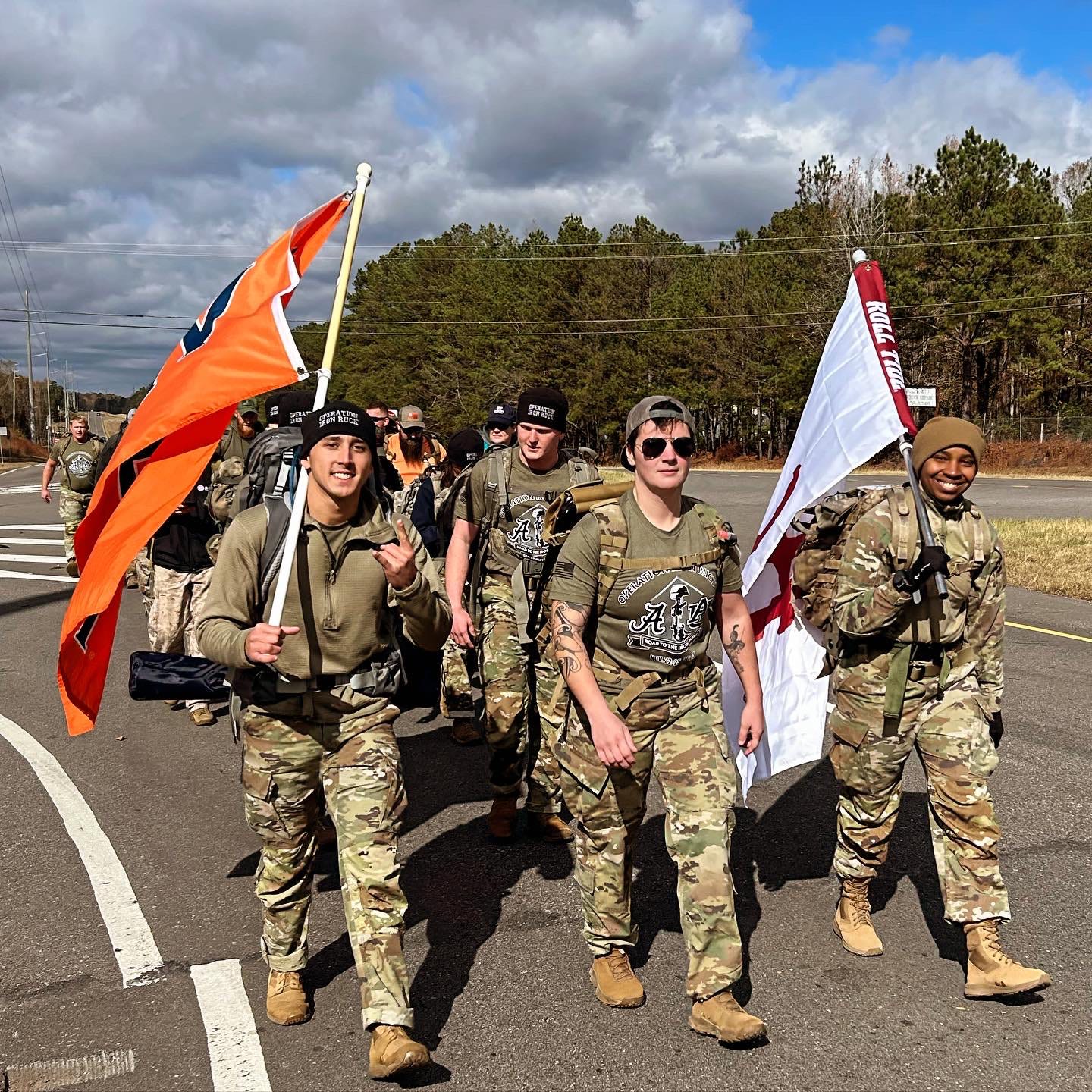 Men dressed in military uniforms with an Auburn University flag and a University of Alabama flag walking down a road.