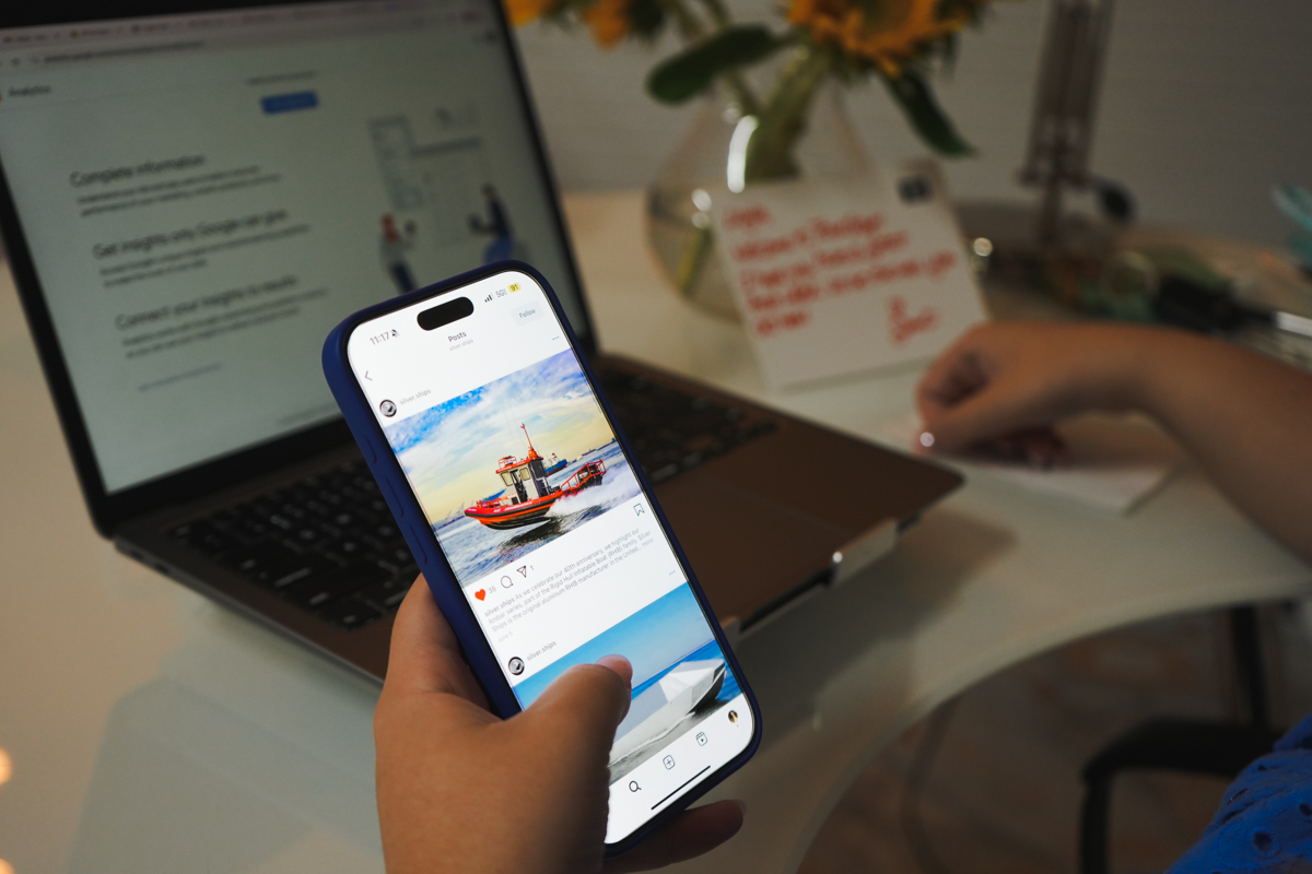 A thumb hovering over Silver Ships instagram feed on an iPhone screen with a laptop on a desk in the background.